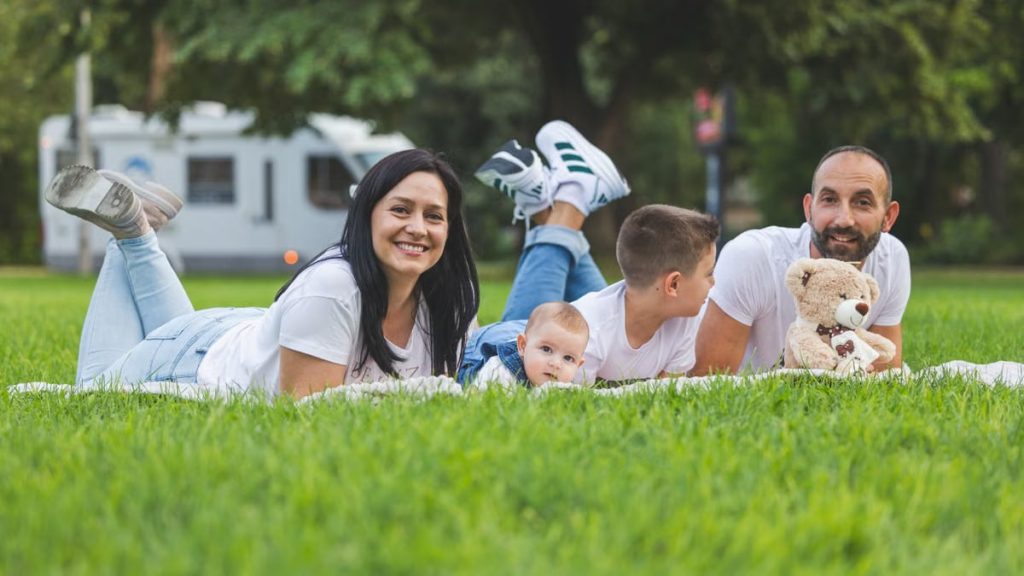 Happy family lying on grass with RV in the background, enjoying a sunny outdoor picnic.